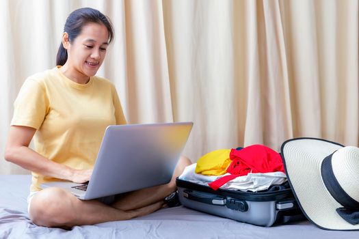 Asian happy woman plan for trip with her friend by laptop on the bed at home. Women with travel bag use notebook computer search for booking hotel frome internet. Holiday summer vacation concept