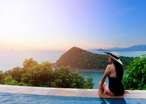 Asian women in black swimsuit and hat siting on side of infinity pool relaxing at sunset time  with sea view during vacation retreat. Women happy and enjoy with holiday summer travel time.