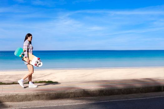 Young women walking on side road near tropical beach with surf skate board. Young girl long hair wear สong sleeve shirt, shot jeans and sneaker walk with surf skate board on the beach road.