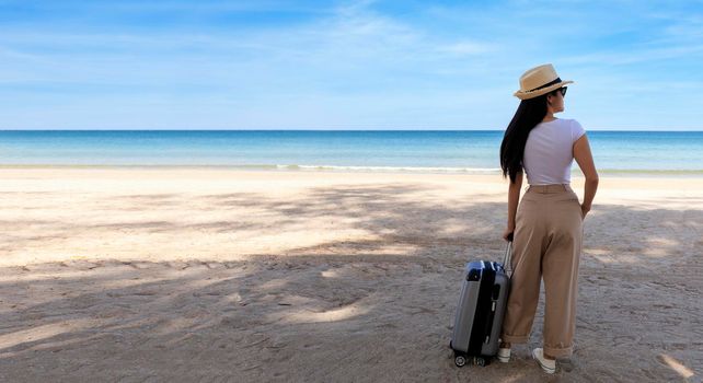 Beautiful young woman wear T-shirt, long pants and straw hat with a suitcase on a tropical beach. Blue sea in the background. Happy travel women standing with travel bag relax on beach. Holiday travel vacation summer