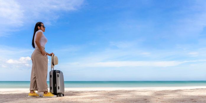 Beautiful young woman wear T-shirt, long pants and straw hat with a suitcase on a tropical beach. Blue sea in the background. Happy travel women standing with travel bag relax on beach. Holiday travel vacation summer