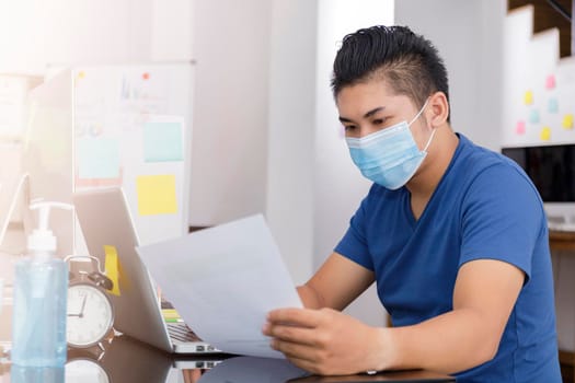 Asian small business start up working at Private Home Office social distancing among Coronavirus outbreak. Businessman wear face mask holding document paper chart .