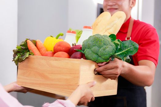 Asian deliver man in red uniform handling wooden crate box of food, fruit, vegetable give to female costumer. Postman and express grocery delivery service
