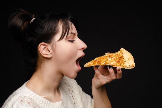 Close-up face of hungry young woman open mouth ready biting appetizing piece of pizza. Casual attractive young female enjoying eating fast food isolated at studio black background side view