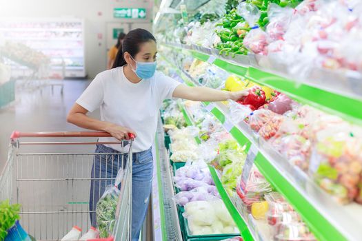 Asian woman wear face mask push shopping cart in supermarket. Girl looking grocery to buy something During coronavirus crisis or covid19 outbreak. Women wearing protective face mask new normal concept