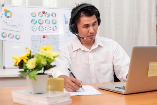 Asian businessman talking with team via video conference writing note on paper. business people using laptop and headset for online meeting. Man using headphone speak with customers. work from home concept.
