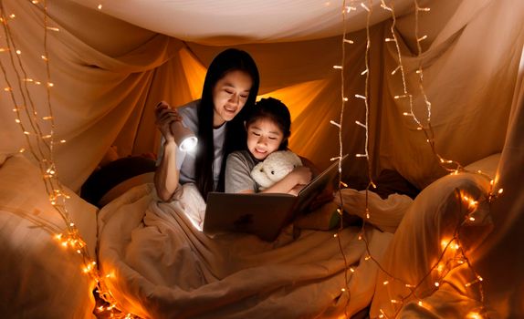 Family concept. elder sister and sister reading book with flashlight together before bedtime. Sister read story book together in bed sheet tent. focus selective little sister. With film grain effect