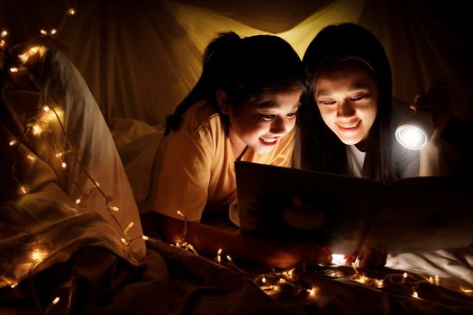 Family concept. Mother and child daughter reading book with flashlight together in children tent before bedtime. Happy mother read story book to her daughter in bed sheet tent
