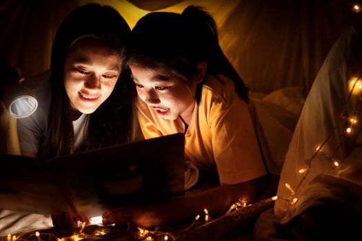 Family concept. Mother and child daughter reading book with flashlight together in children tent before bedtime. Happy mother read story book to her daughter in bed sheet tent
