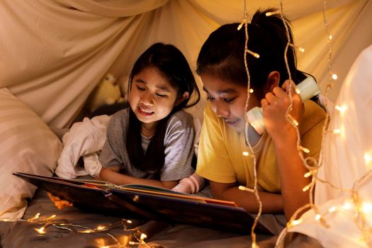 Family concept. elder sister and sister reading book with flashlight together in children tent before bedtime. Happy elder sister read story book to her sister in bed sheet tent
