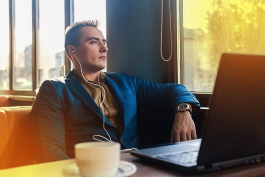 A business serious man businessman a stylish of Caucasian appearance, works in a laptop or computer, sitting at a table by the window in a cafe and listening to music on headphones.