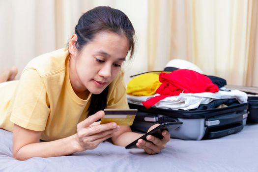 Asian happy woman plan for trip by mobile phone and credit card on the bed at home. Women with travel bag use smart phone and credit card pay booking hotel. Holiday summer concept