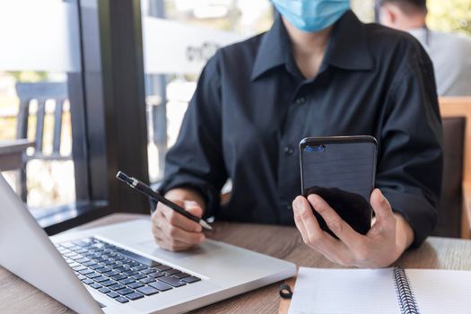 Young woman with protective face mask sitting in coffee shop at wood table using smartphone.and laptop. Girl browsing internet, chatting, blogging. Female holding phone and looking on screen