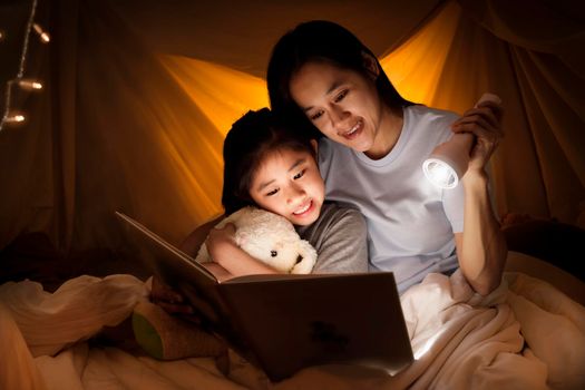 Family concept. Mother and child daughter reading book with flashlight together in children tent before bedtime. Happy mother read story book to her daughter in bed sheet tent
