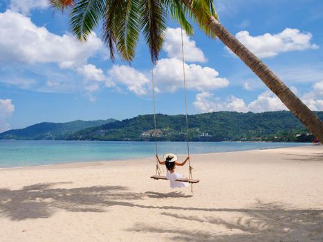 Phuket, Thailand. Tropical beach paradise with beach swing with girl in white shirt. Women relax on swing under coconut palm tree at beautiful tropical beach White sand. Luxury beach scene vacation summer holiday concept.