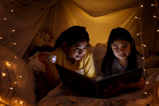 Family concept. elder sister and sister reading book with flashlight together in children tent before bedtime. Happy elder sister read story book to her sister in bed sheet tent