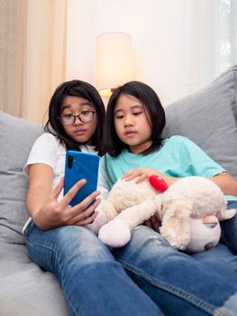 Happy siblings children sitting on sofa in living room speak with parent on mobile phone together, smiling elder girl showing cute little sister video call by smartphone speak with family.