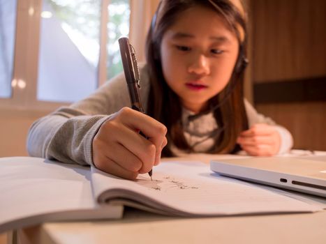 Close up hand of  Asian girl in headphones sit at desk study online on laptop. Kid wear headset handwriting in notebook learning using internet lessons on quarantine. Girl student learning virtual internet online class from school due to covid pandemic.