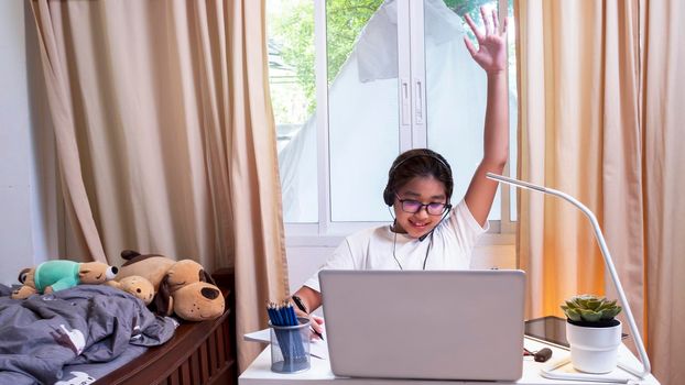Asian girl in headphones sit at desk study online on laptop. Kid wear headset handwriting in notebook learning using internet lessons on quarantine. Girl student learning virtual internet online class from school due to covid pandemic.