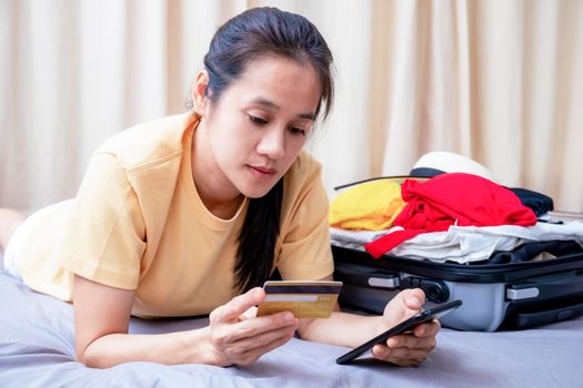 Asian happy woman plan for trip by mobile phone and credit card on the bed at home. Women with travel bag use smart phone and credit card pay booking hotel. Holiday summer concept