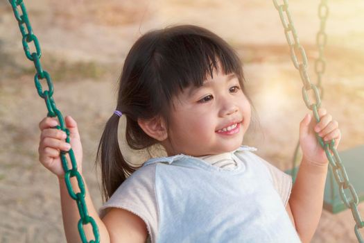 Close up cute little girl having fun on play swing in playground on warm and sunny day outdoors. Asian girl happy enjoy to play swing at playground