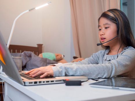  Asian girl in headphones sit at desk study online on laptop. Kid wear headset typing keyboard notebook learning using internet lessons on quarantine. Girl student learning virtual internet online class from school due to covid pandemic.