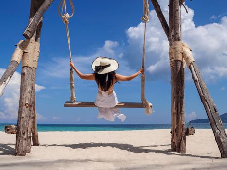 Happy young woman enjoy and sway the wooden swing over the sea beach, blue sky and cloud. Tourist women relax and enjoy with wood swing on the beach Phuket, Thailand. Summer holiday vacition concept.
