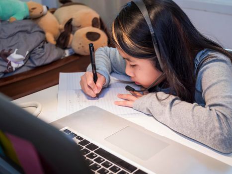  Asian girl in headphones sit at desk study online on laptop. Kid wear headset handwriting in notebook learning using internet lessons on quarantine. Girl student learning virtual internet online class from school due to covid pandemic.