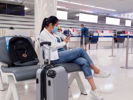 Asian traveler women wear face masks sitting social distancing chairs from corona virus pandemic at the airport. People travel with new normal lifestyle concepts. Young women sitting and use mobile phone at airport