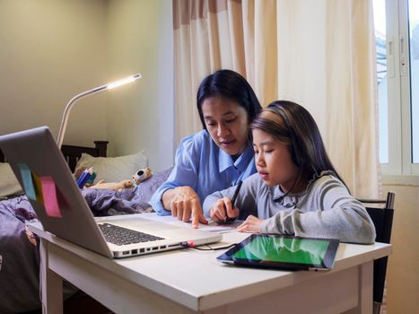 Asian young little girl using pencil to do homework with her mother. Student kid writing  homework book. Girl use computer to study at home. Education homeschool  concept.