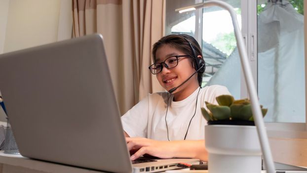  Asian girl in headphones sit at desk study online on laptop. Kid wear headset handwriting in notebook learning using internet lessons on quarantine. Girl student learning virtual internet online class from school due to covid pandemic.