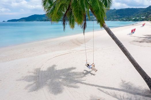 Phuket, Thailand. Tropical beach paradise with beach swing with girl in white shirt. Women relax on swing under coconut palm tree at beautiful tropical beach White sand. Luxury beach scene vacation summer holiday concept.