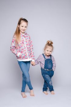 Front view of two pretty sisters keeping hands, looking at camera and smiling on isolated background in studio. Happy kids wearing stylish outfits posing together. Concept of friendship and fashion.
