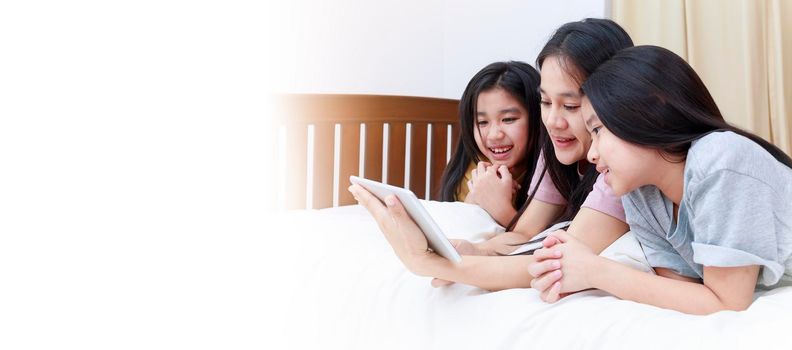 Happy Asian family enjoy and relax on bed in bedroom. mother and daughters enjoy using tablet together on bed.  Family concept. With copy space banner.