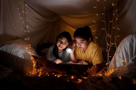 Family concept. elder sister and sister reading book with flashlight together before bedtime. Sister read story book together in bed sheet tent. focus selective little sister. With film grain effect