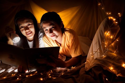 Family concept. elder sister and sister reading book with flashlight together before bedtime. Sister read story book together in bed sheet tent. focus selective little sister. With film grain effect