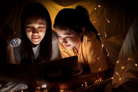 Family concept. Mother and child daughter reading book with flashlight together in children tent before bedtime. Happy mother read story book to her daughter in bed sheet tent
