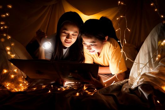 Family concept. Mother and child daughter reading book with flashlight together in children tent before bedtime. Happy mother read story book to her daughter in bed sheet tent
