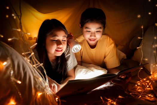 Family concept. elder sister and sister reading book with flashlight together before bedtime. Sister read story book together in bed sheet tent. focus selective little sister. With film grain effect