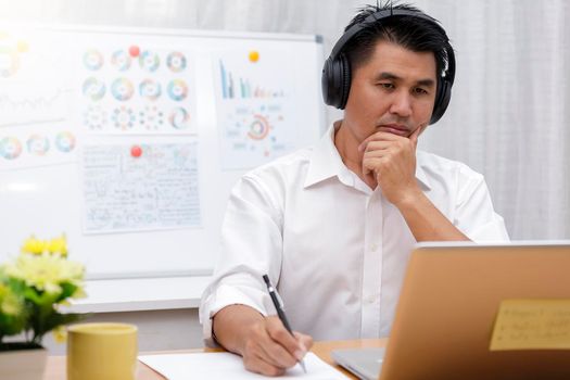 Asian businessman talking with team via video conference writing note on paper. business people using laptop and headset for online meeting. Man using headphone speak with customers. work from home concept.