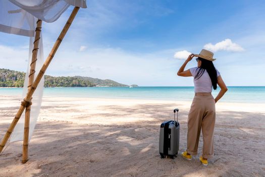 Beautiful young woman wear T-shirt, long pants and straw hat with a suitcase on a tropical beach. Blue sea in the background. Happy travel women standing with travel bag relax on beach. Holiday travel vacation summer
