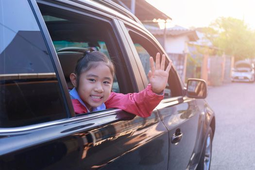 On the way to school, the little girl stretched out his hand from the car window, laughing and smiling. Asian little girl smiling  and waving hand out the car. Children relax with street view from the car. Family in car concept.