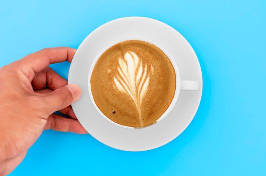 Top view hand man holding  white coffee coasters with Cappuccino coffee and creamy shape in white cup. Hand man hold coffee cup on blue table in coffee shop.