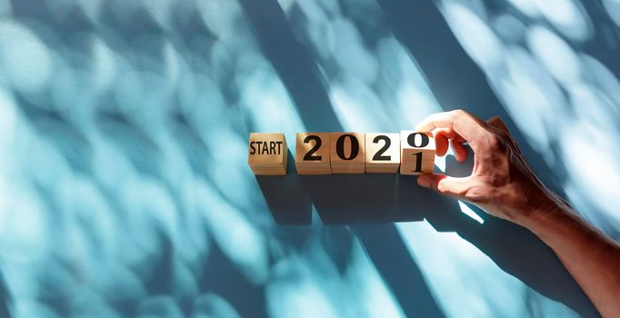 Hand holding wooden cube with flip over block start 2020 to 2021 word on blue and shadow background. Top view hand flip wood block with number text start 2020, 2021 with copy space