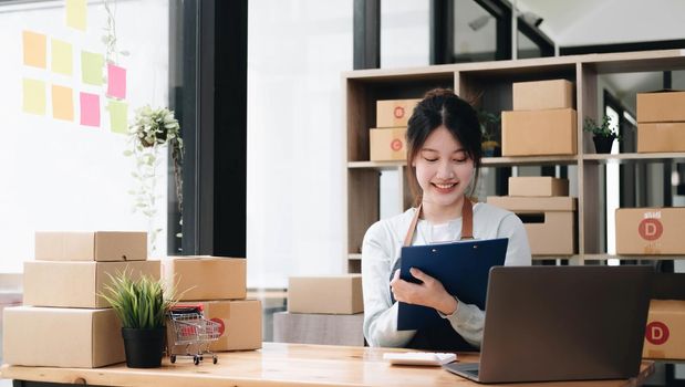 A portrait of a young Asian woman, e-commerce employee sitting in the office full of packages in the background write note of orders and a calculator, for SME busines e-commerce and delivery business