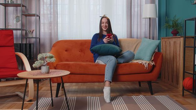 Happy excited joyful stylish teen girl in sweater use mobile cell phone typing browsing say wow yes found out great big win news doing winner gesture. Young adult woman at home in living room on sofa