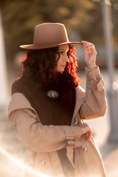 Outdoor fashion portrait of young elegant fashionable brunette woman, model in stylish hat, choker and light raincoat posing at sunset in European city