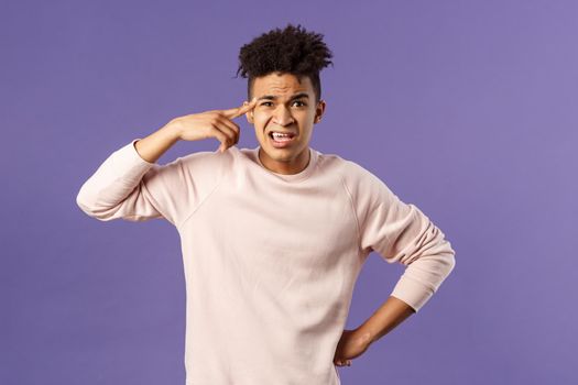 Portrait of angry, annoyed young man scolding someone from being stupid and crazy, rolling index finger over temple staring outraged and irritated camera, standing bothered purple background.