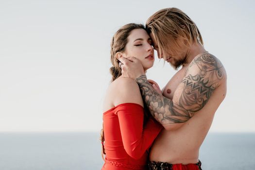 Portrait of tattooed man and sensual young brunette woman in red dress, happily dancing outdoors isolated on blurry sea background with natural bokeh in soft warm sunset backlight. Selective focus