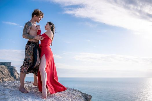 Portrait of tattooed man and sensual young brunette woman in red dress, happily dancing outdoors isolated on blurry sea background with natural bokeh in soft warm sunset backlight. Selective focus